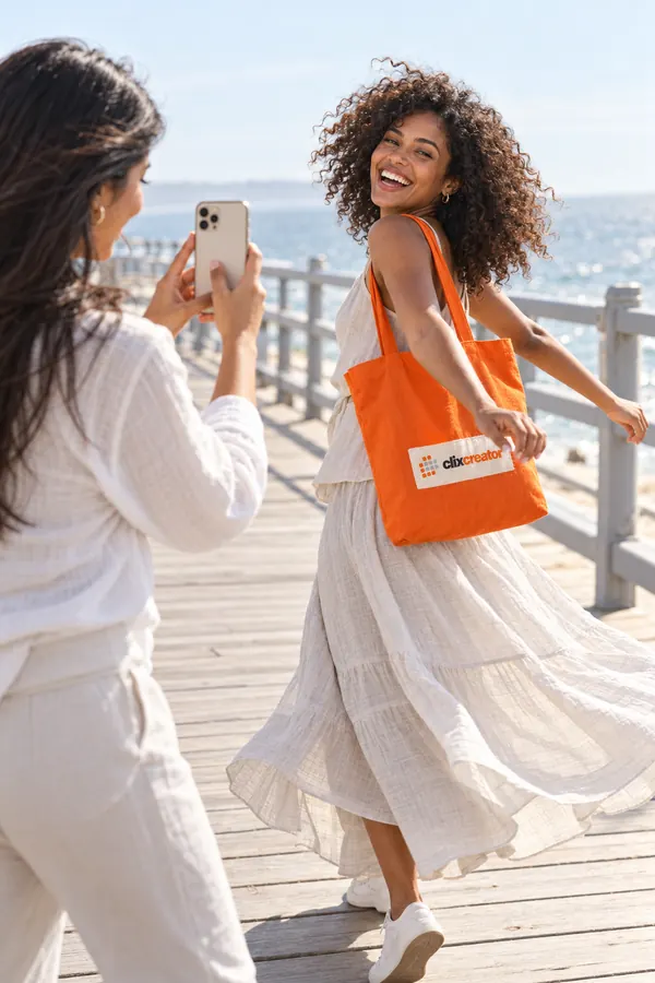 Young woman filming her friend spinning on a sunny seaside boardwalk, the friend carries an orange Clix Creator tote bag